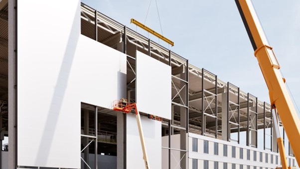 A construction site showing a crane installing a prefabricated wall panel on a battery plant, with workers on an elevated platform ensuring precision.