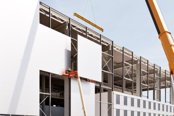 A construction site showing a crane installing a prefabricated wall panel on a battery plant, with workers on an elevated platform ensuring precision.